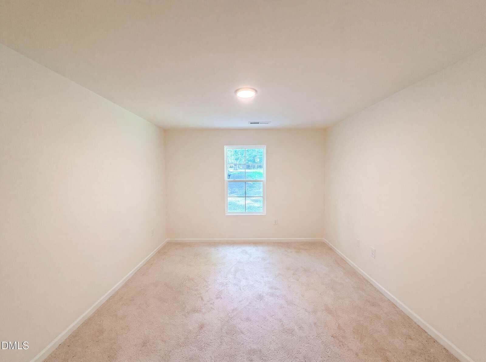 A bright, empty room with cream-colored walls, beige carpet, a single ceiling light, and a window centered on the far wall letting in natural light.