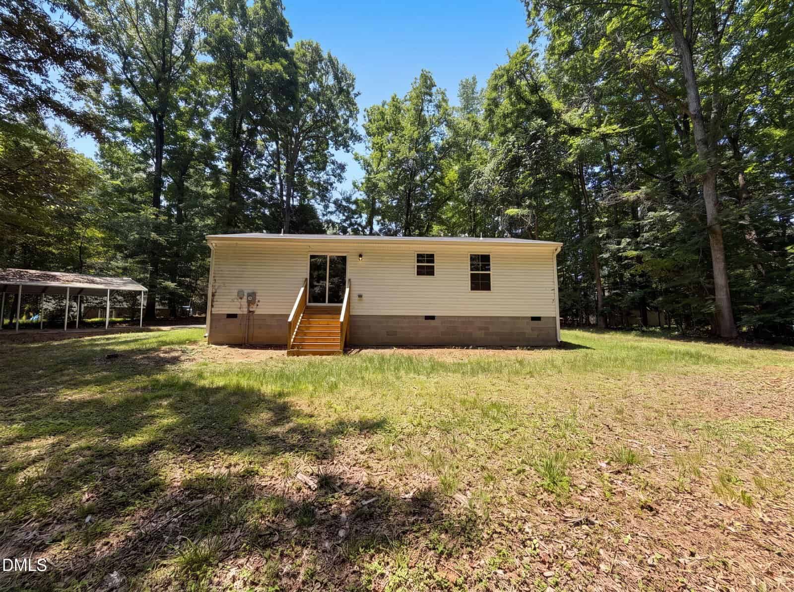 A small, single-story house with beige siding and a few windows, sits in a grassy yard surrounded by tall trees. Wooden steps lead up to a glass back door. A carport is visible on the left side.