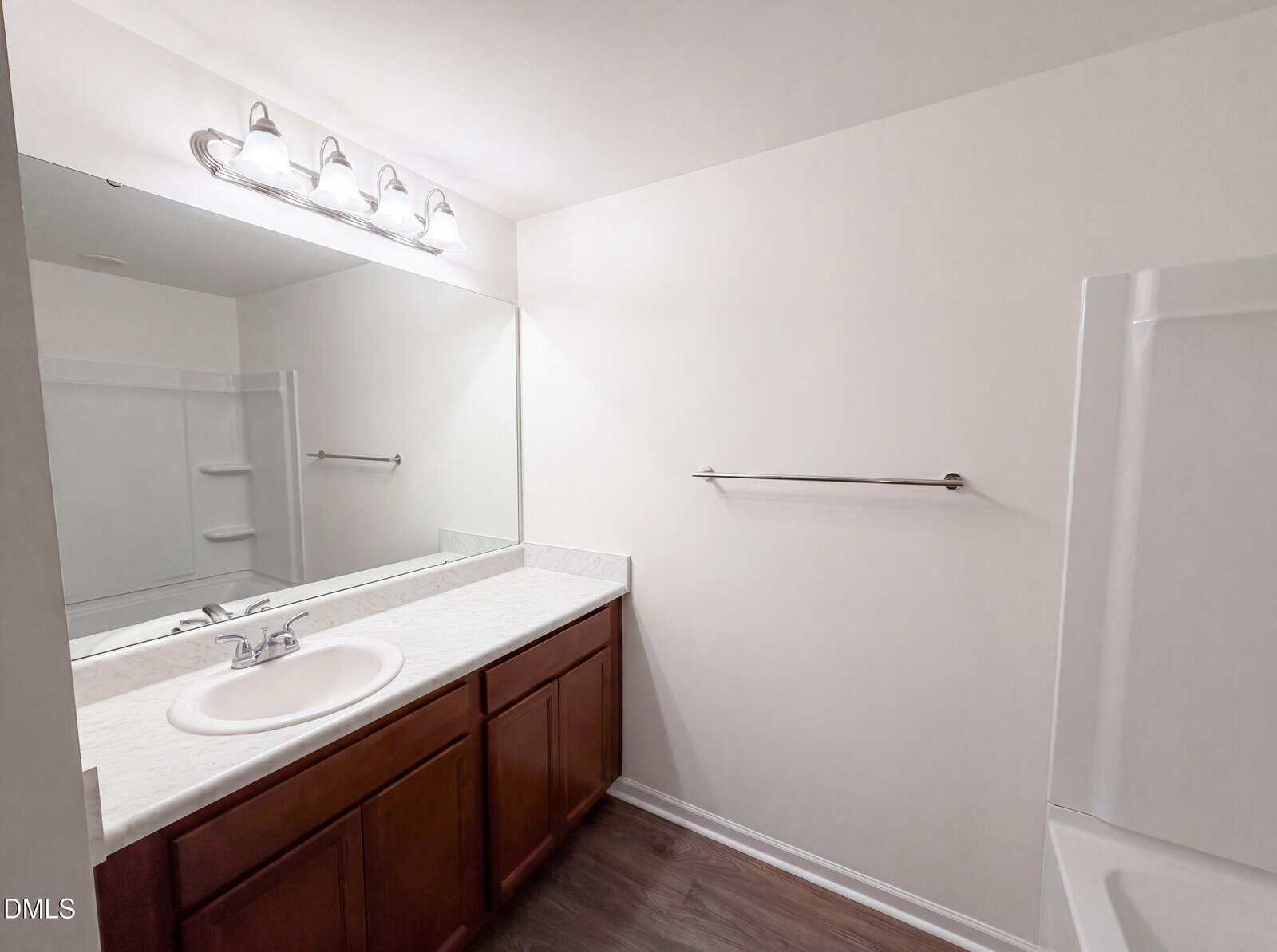 A bathroom with a large mirror, single sink, wooden vanity, overhead lights, towel bar, and a bathtub with built-in shelves. The walls are white and the floor is dark wood.