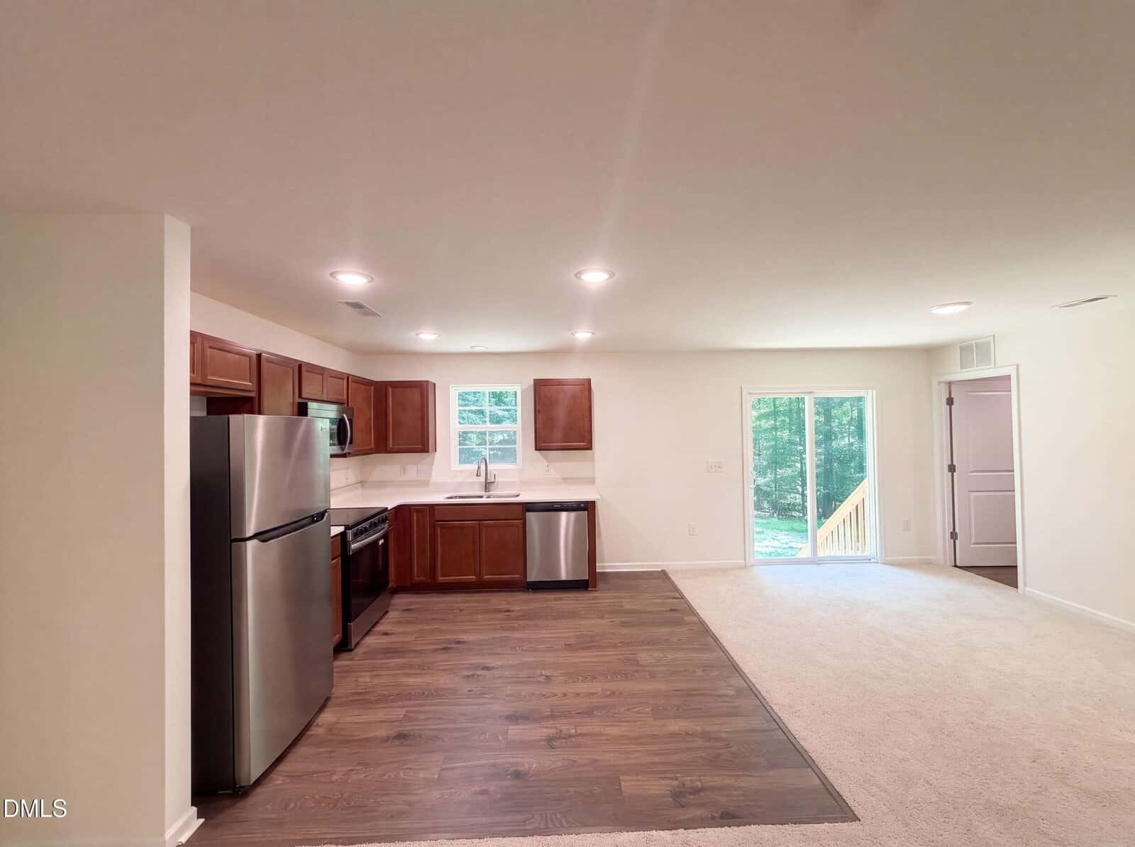 Open kitchen with stainless steel appliances and wooden cabinets, adjacent to a bright living area with sliding glass doors leading outside. Hardwood and carpet flooring are visible.