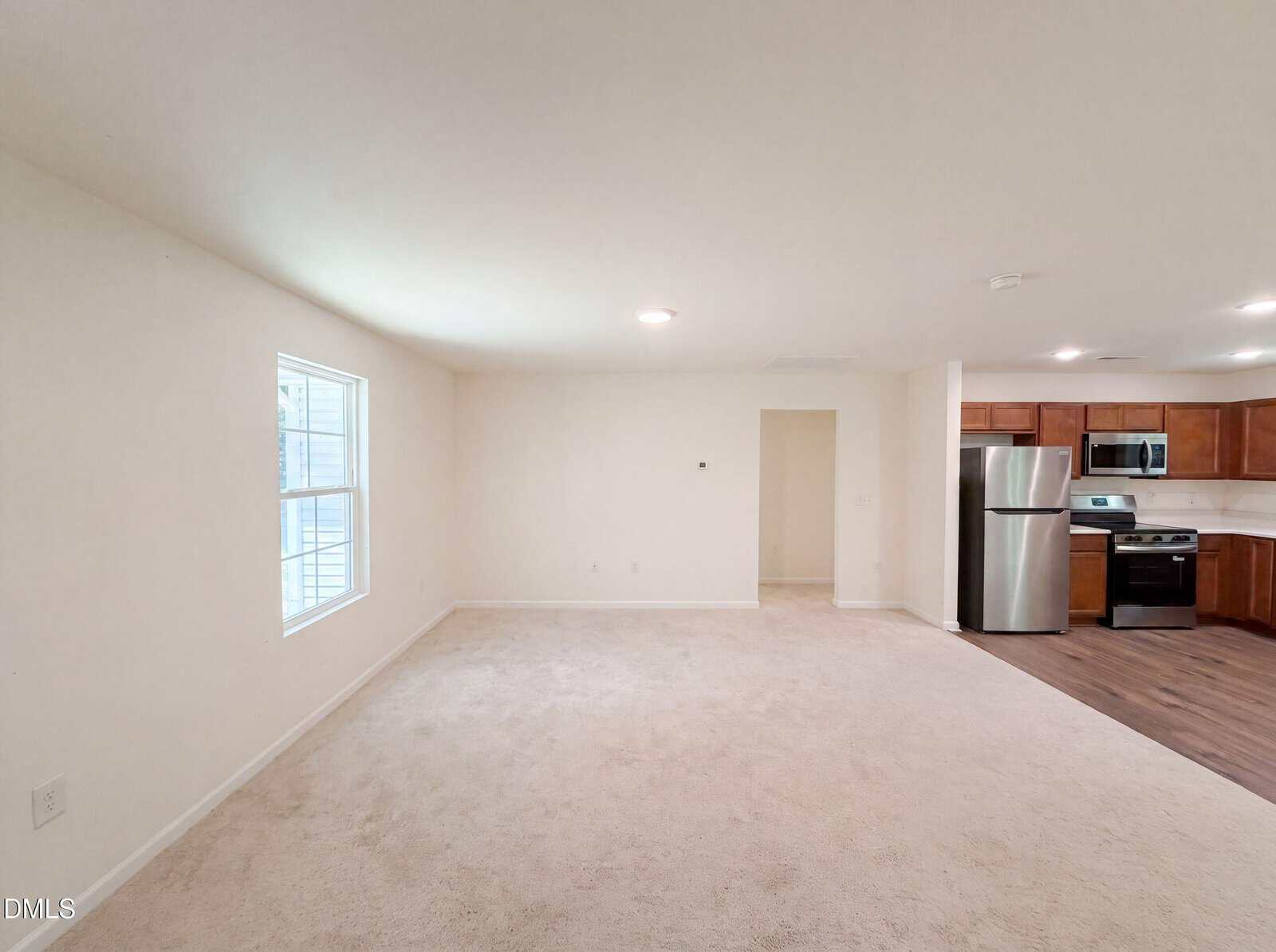 Bright, empty living room with beige carpet, white walls, a window on the left, and an open view into a modern kitchen with wooden cabinets and stainless steel appliances on the right.