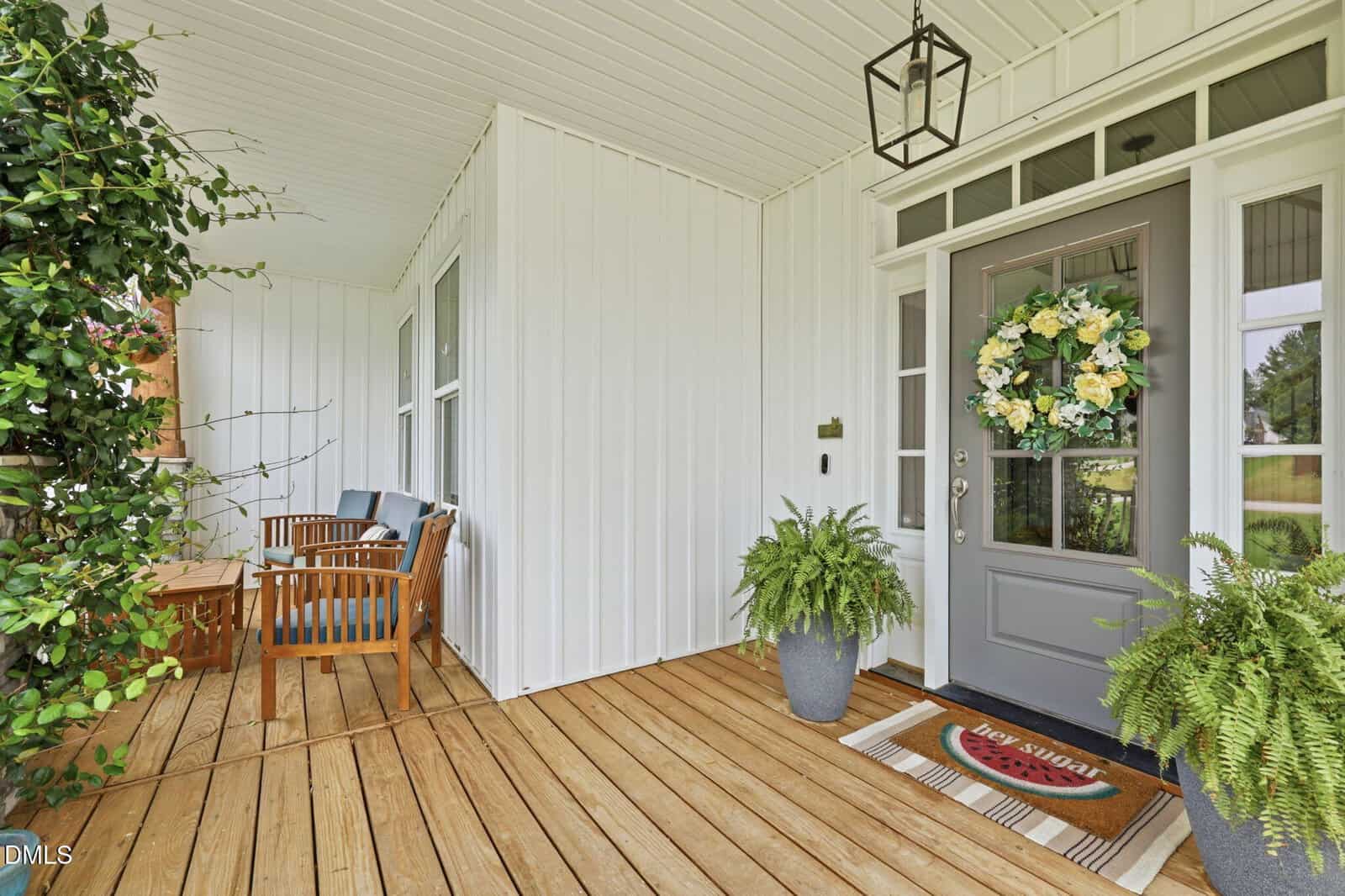 A cozy porch with wooden flooring, a gray front door with a floral wreath, two potted plants, a doormat, and a seating area with blue cushions and a wooden table.
