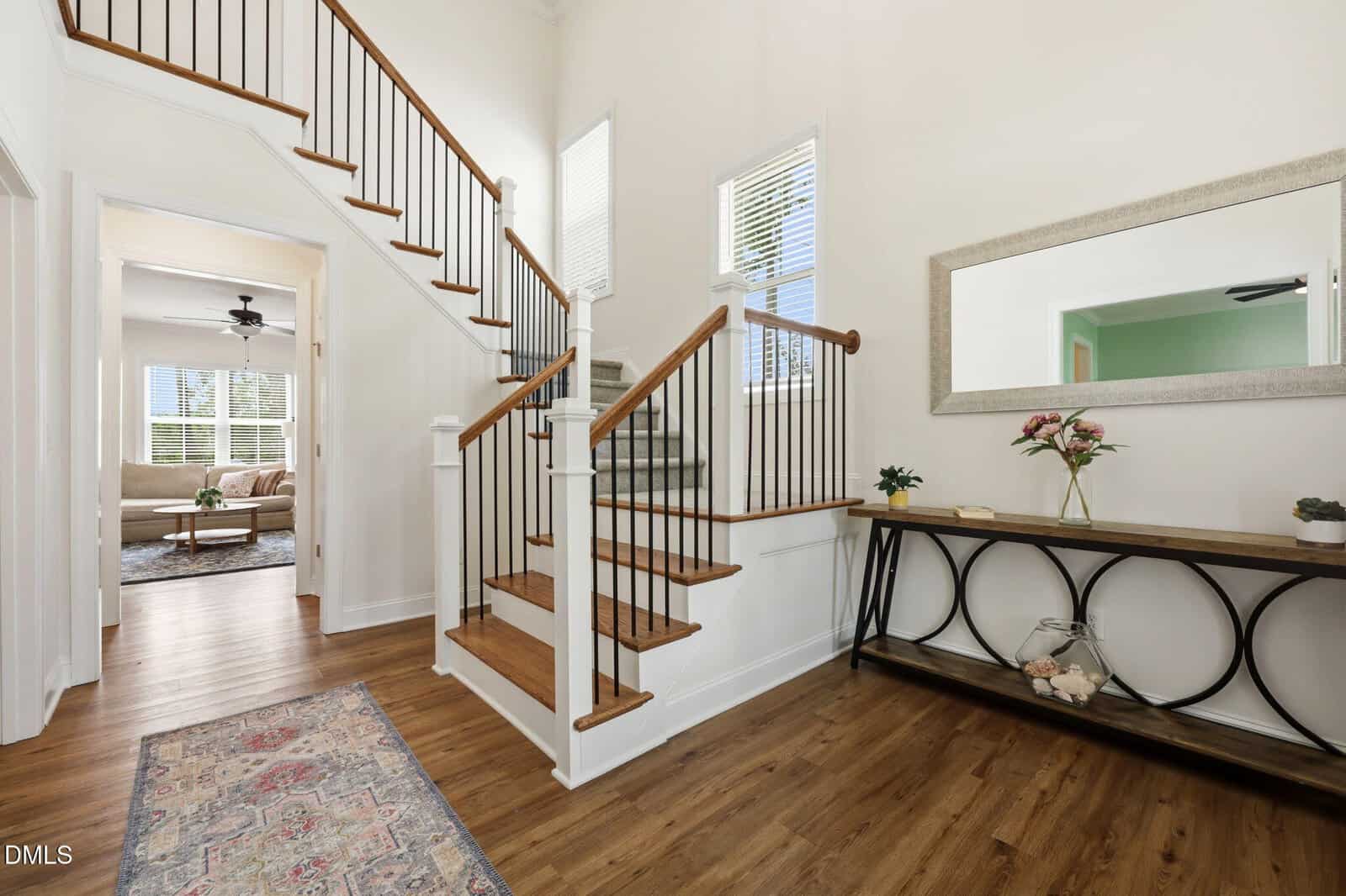 Bright entryway with wooden stairs and white walls, featuring a console table with flowers and decor, a large mirror, and a view into a cozy living room with a sofa, rug, and ceiling fan.