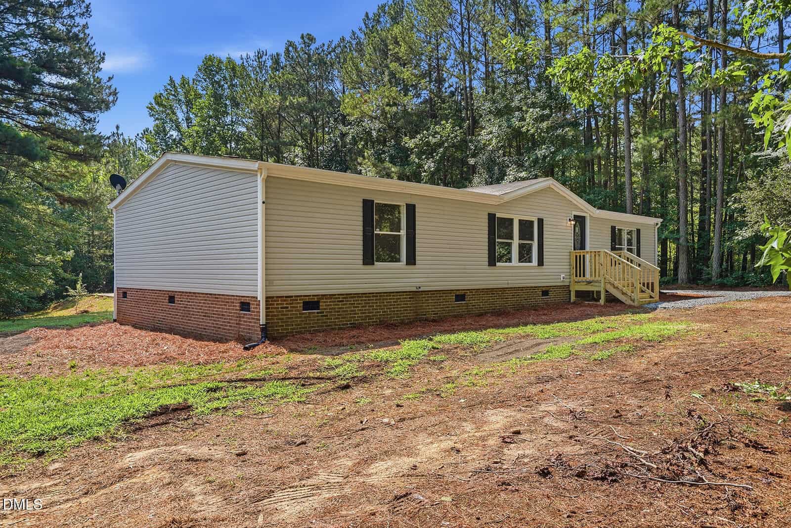 A beige manufactured home with a brick foundation sits amid tall pine trees. The home has several windows, a small wooden front porch with steps, and is surrounded by grassy and mulched areas under a clear blue sky.