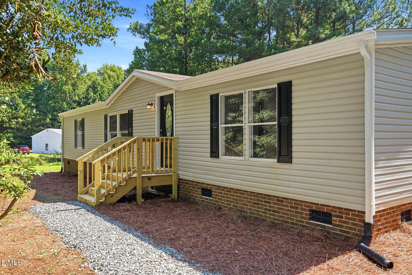 Single-story beige manufactured home with brick foundation, black shutters, and a small wooden porch with steps. Gravel walkway and pine straw landscaping, surrounded by trees and greenery under a clear sky.