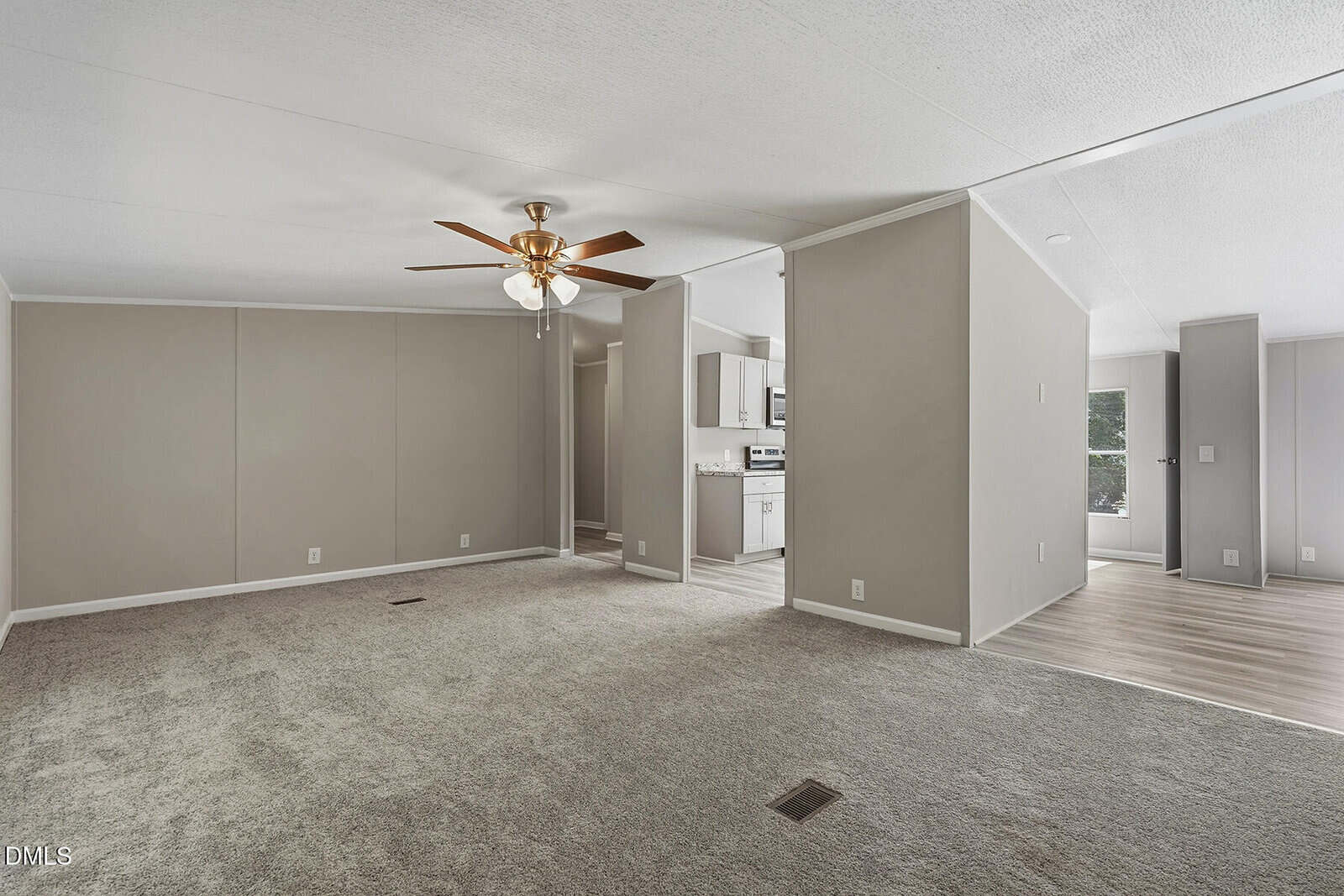 Spacious living area with gray carpet, beige walls, and ceiling fan, leading to an open kitchen with white cabinets and stainless steel appliances. Light wood flooring is visible in the kitchen and adjacent space.