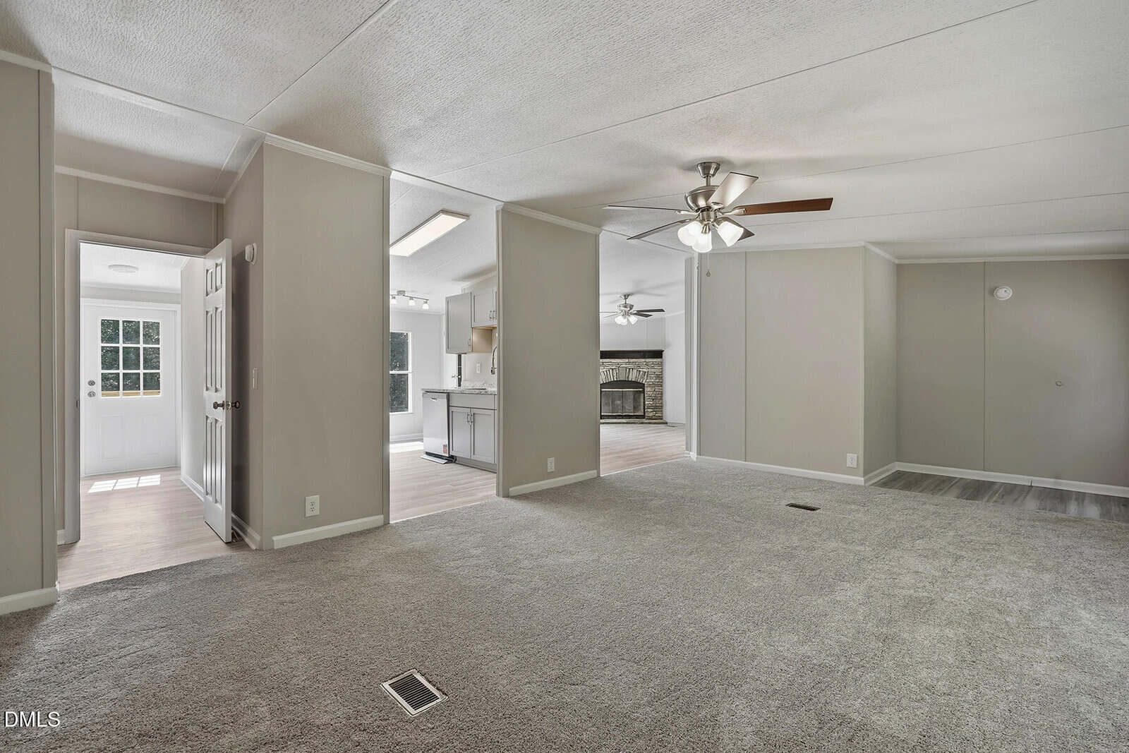 A spacious living area with light gray carpet, neutral walls, ceiling fans, and an open layout leading to a kitchen with wood flooring. Natural light enters through a glass door and windows.
