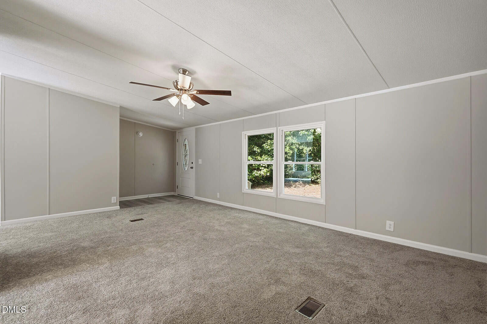A spacious living room with beige carpet, light gray walls, a ceiling fan with lights, two windows showing greenery outside, and a closed front door in the background. The room is empty of furniture.