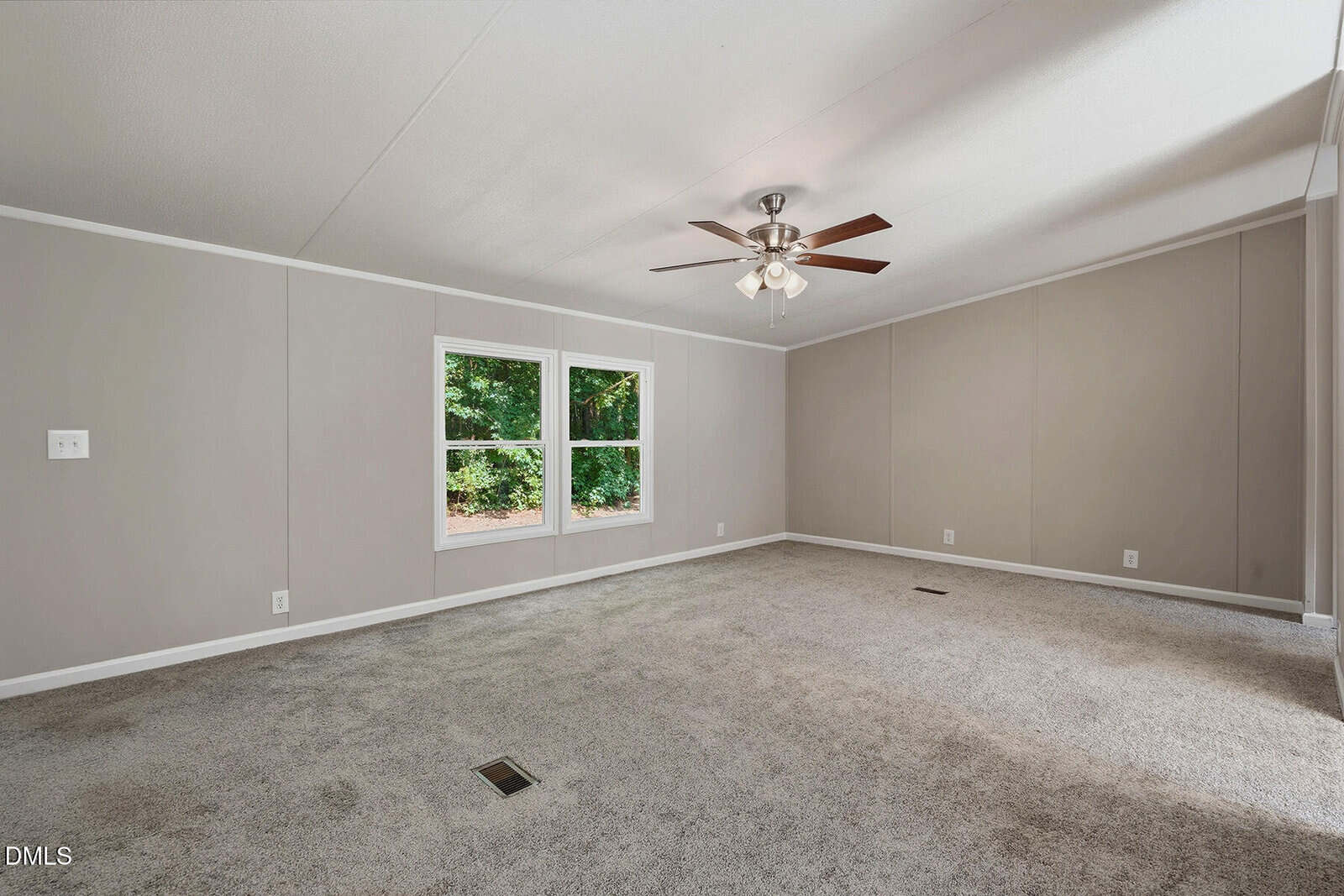 Empty carpeted room with beige walls, white trim, large windows showing greenery outside, and a ceiling fan with lights. Sunlight enters through the windows, illuminating part of the floor. No furniture or decorations are present.