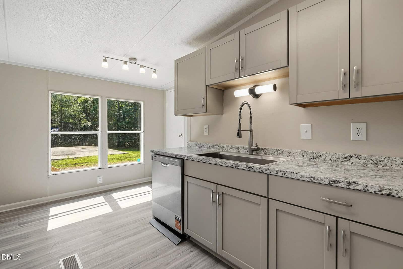 Bright kitchen with large window, modern faucet, white cabinets, gray countertop, and stainless steel sink. Sunlight streams onto the light wood floor. A light fixture hangs from the ceiling near the window.