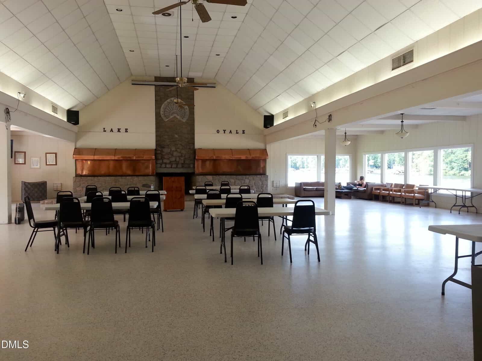 A spacious hall with white walls and ceiling, several rectangular tables and black chairs arranged in the center, a stone fireplace at the far end, and windows lining the right wall letting in natural light.