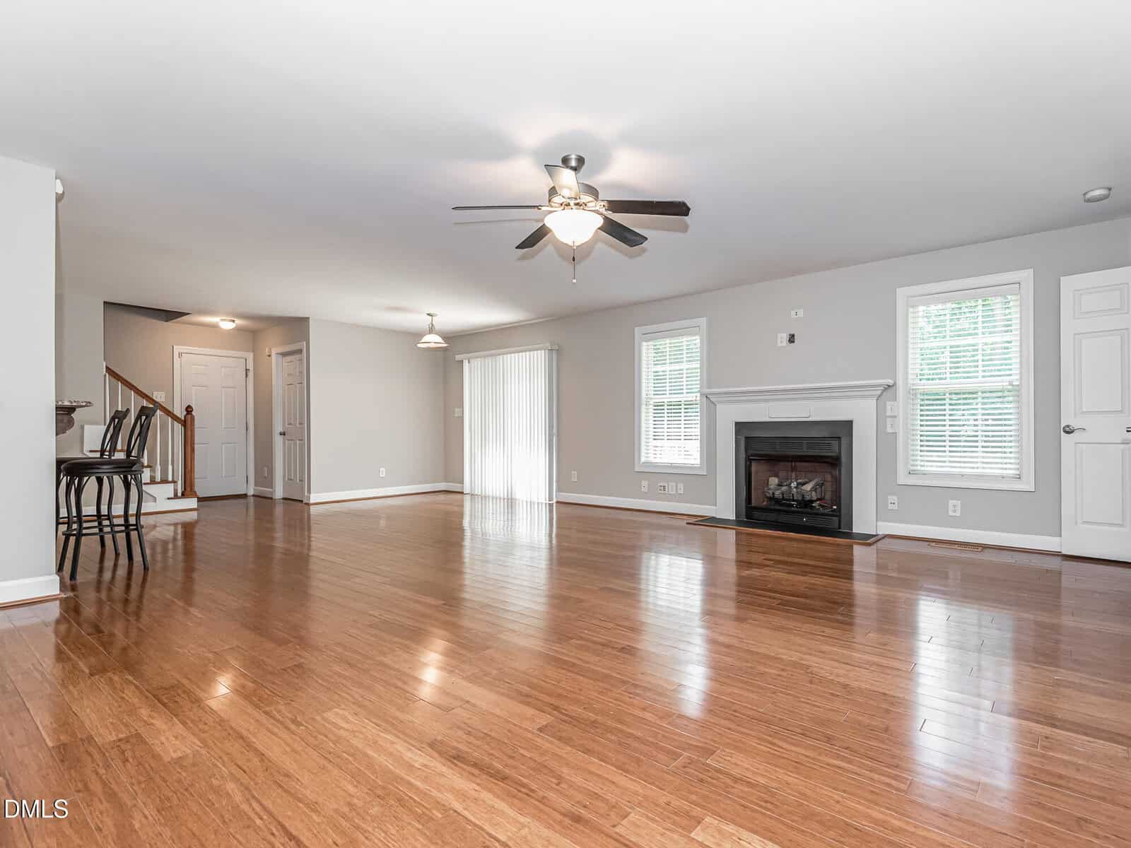 Spacious living room with wooden floors, white walls, a fireplace, ceiling fans, large windows, a staircase, and a breakfast bar with two stools near the kitchen.
