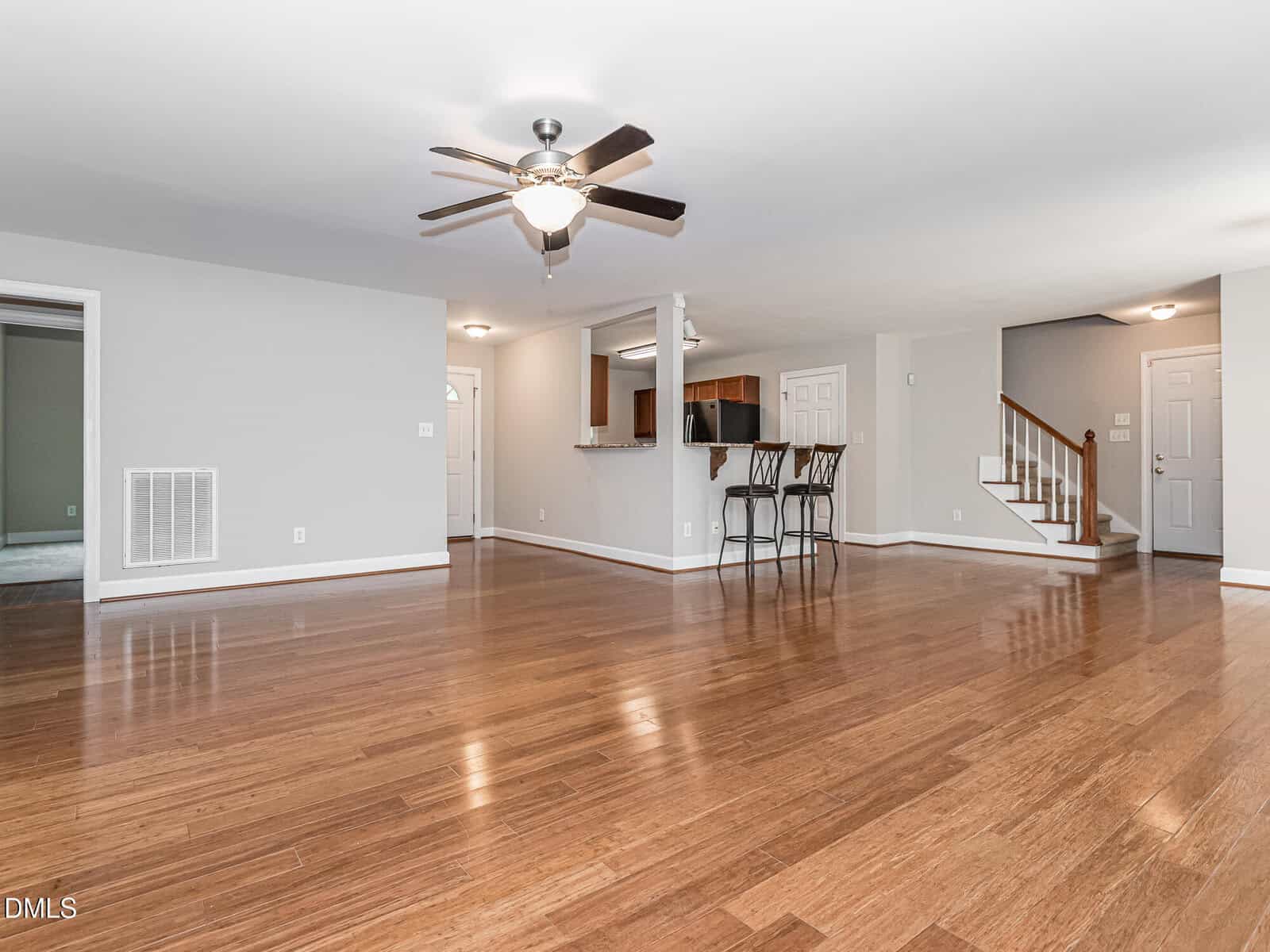Spacious empty living room with wood floors, ceiling fan, open kitchen with bar stools, white walls, and staircase leading upstairs.