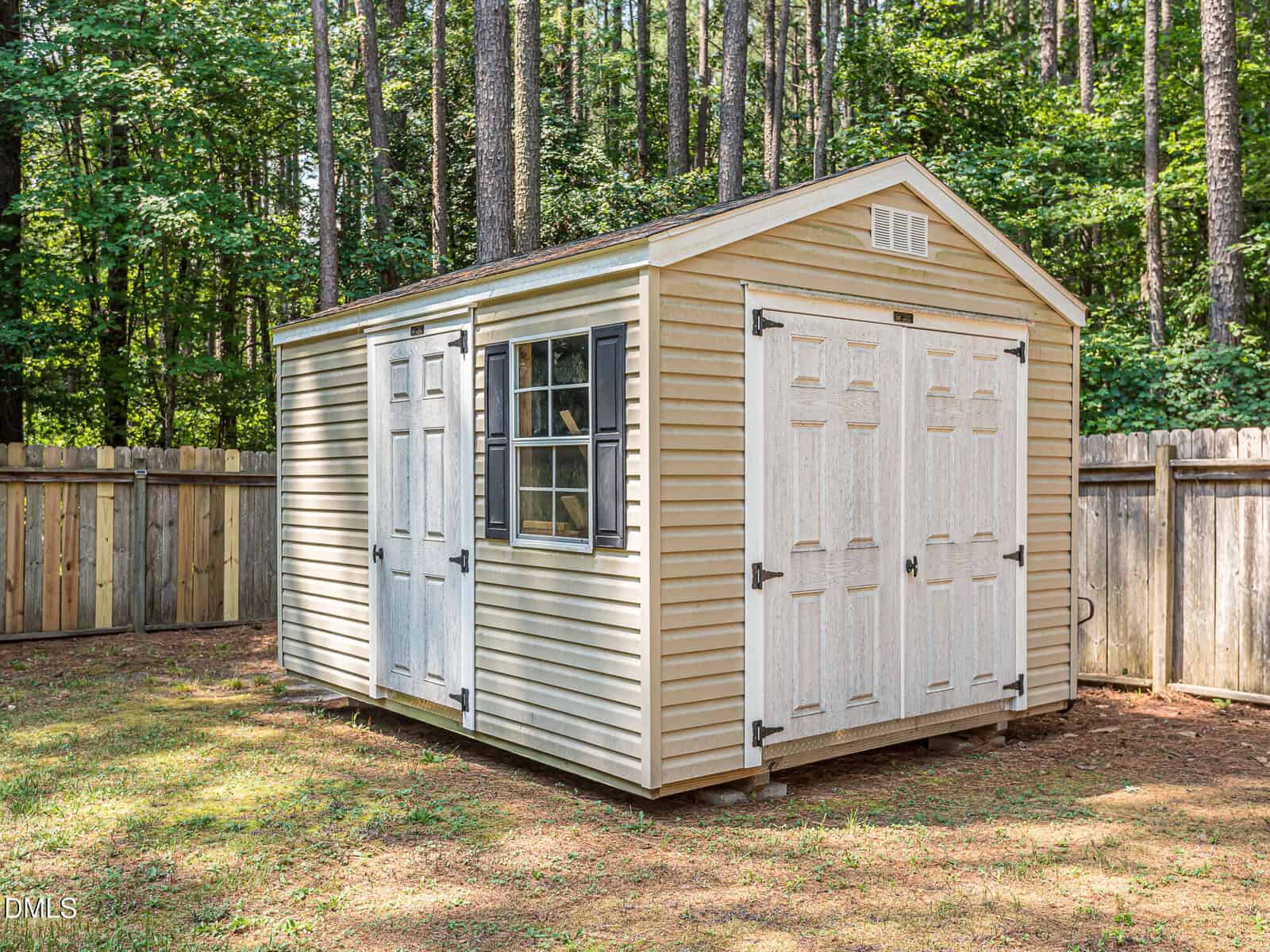 A beige storage shed with white double doors and a window stands in a fenced backyard surrounded by trees and greenery.