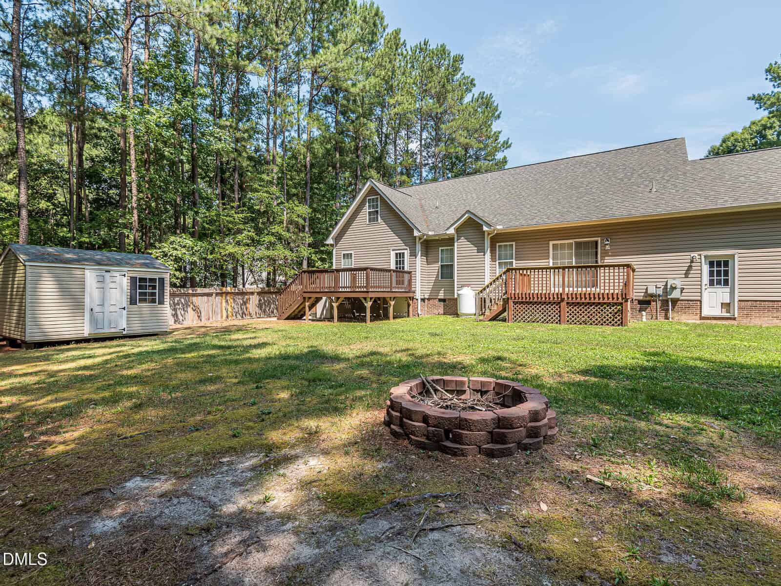 A spacious backyard with a fire pit in the foreground, a wooden deck attached to a beige house, a small storage shed, and surrounded by tall trees under a clear sky.