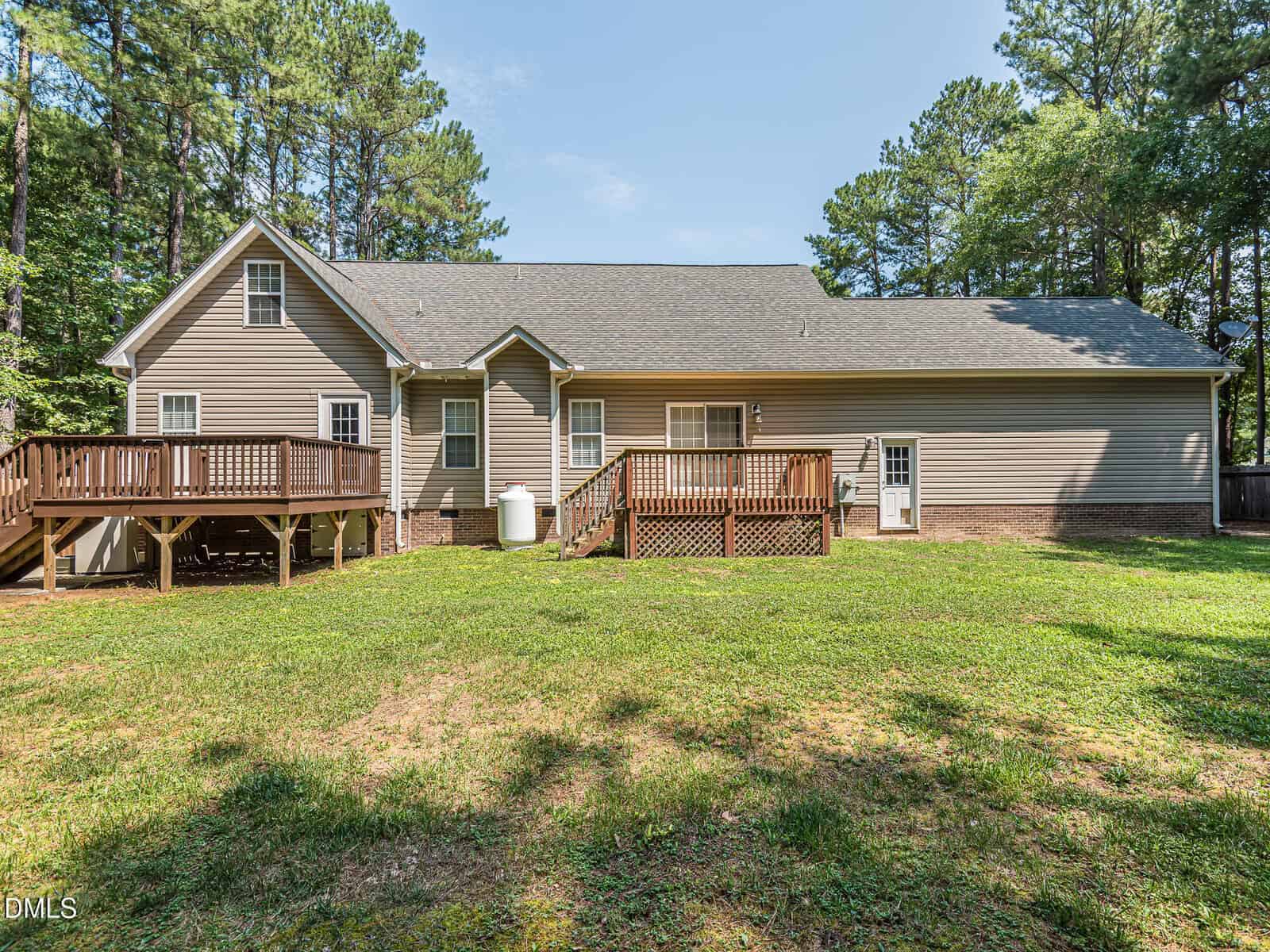Single-story house with beige siding and a gray roof, set in a grassy backyard. Two wooden decks are attached at the back, surrounded by tall trees under a clear blue sky.