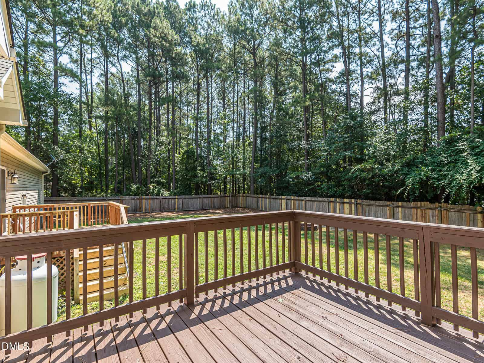 A wooden deck overlooks a fenced backyard with grass and tall pine trees beyond the fence. The deck has a railing and steps leading down to the yard. The sky is clear and sunny.