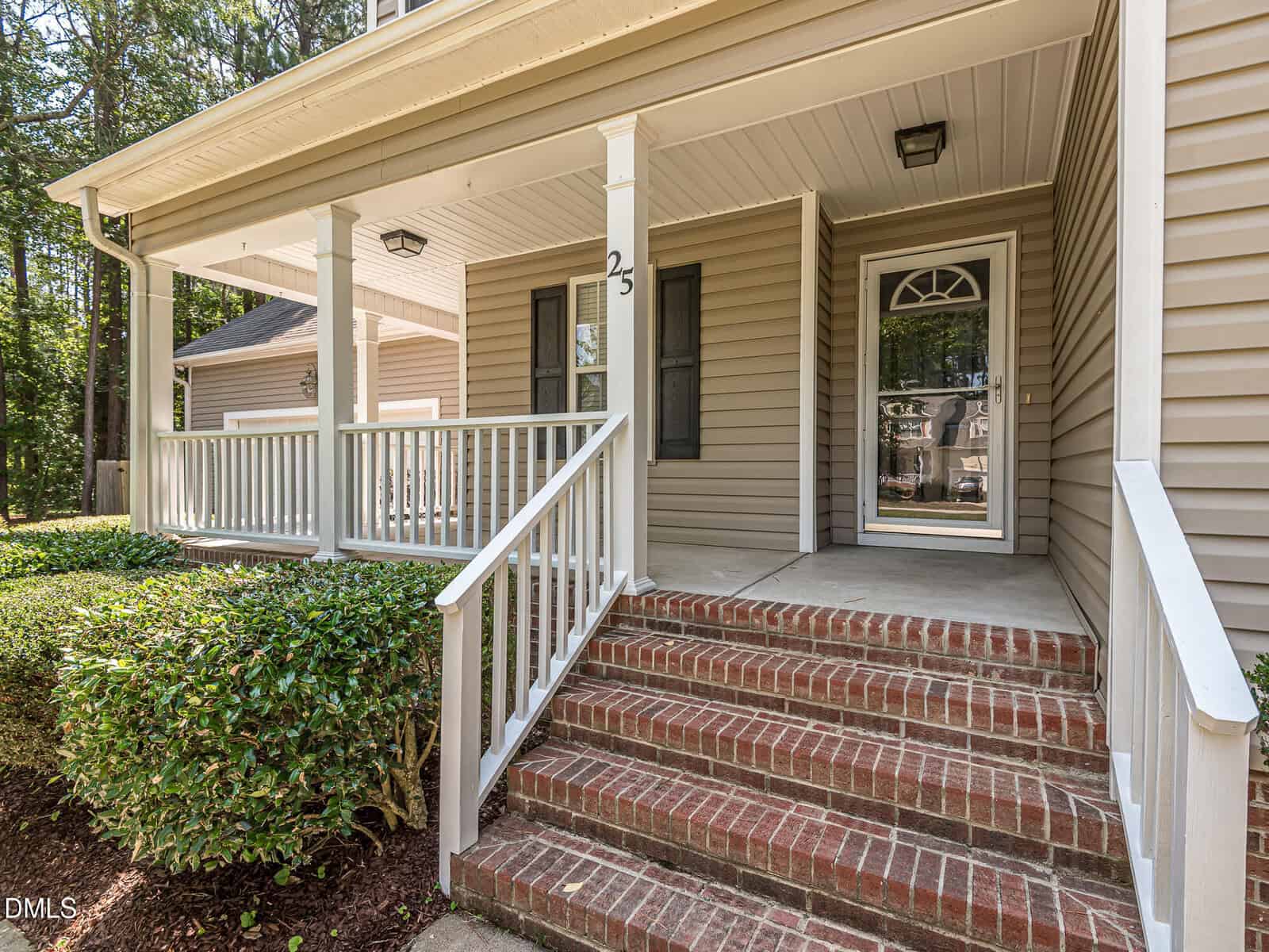 A brick staircase with white railings leads to a covered porch of a beige house with a glass front door and white trim. Bushes line the walkway, and trees are visible in the background.