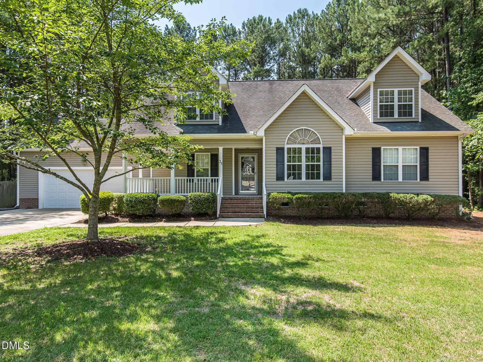 A beige two-story suburban house at 25 Shellcross Court in Youngsville with white trim, a front porch, gabled roof, attached garage, and a well-kept lawn surrounded by trees and greenery on a sunny day.