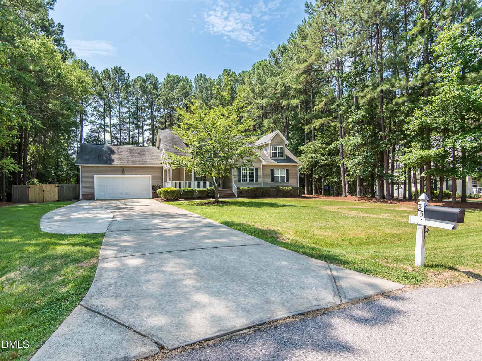 A light-colored suburban house with a two-car garage, a wide driveway, and a mailbox marked "23." The house is surrounded by green grass, trees, and a wooded area in the background.