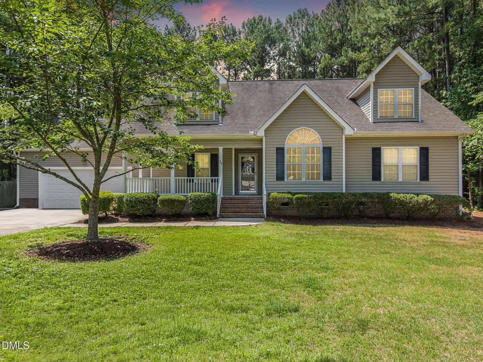 A two-story suburban house with beige siding, black shutters, and a front porch, located at 25 Shellcross Court in Youngsville. The yard is grassy with a tree and trimmed bushes. The driveway leads to a two-car garage, while tall trees line the background.