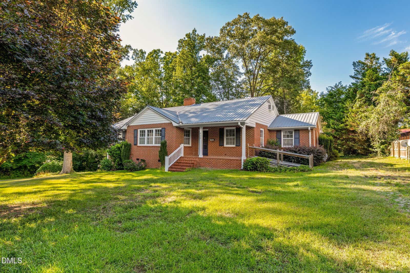 A red brick house with a metal roof, front porch, and a ramp, surrounded by green grass, large trees, and shrubs under a blue sky.