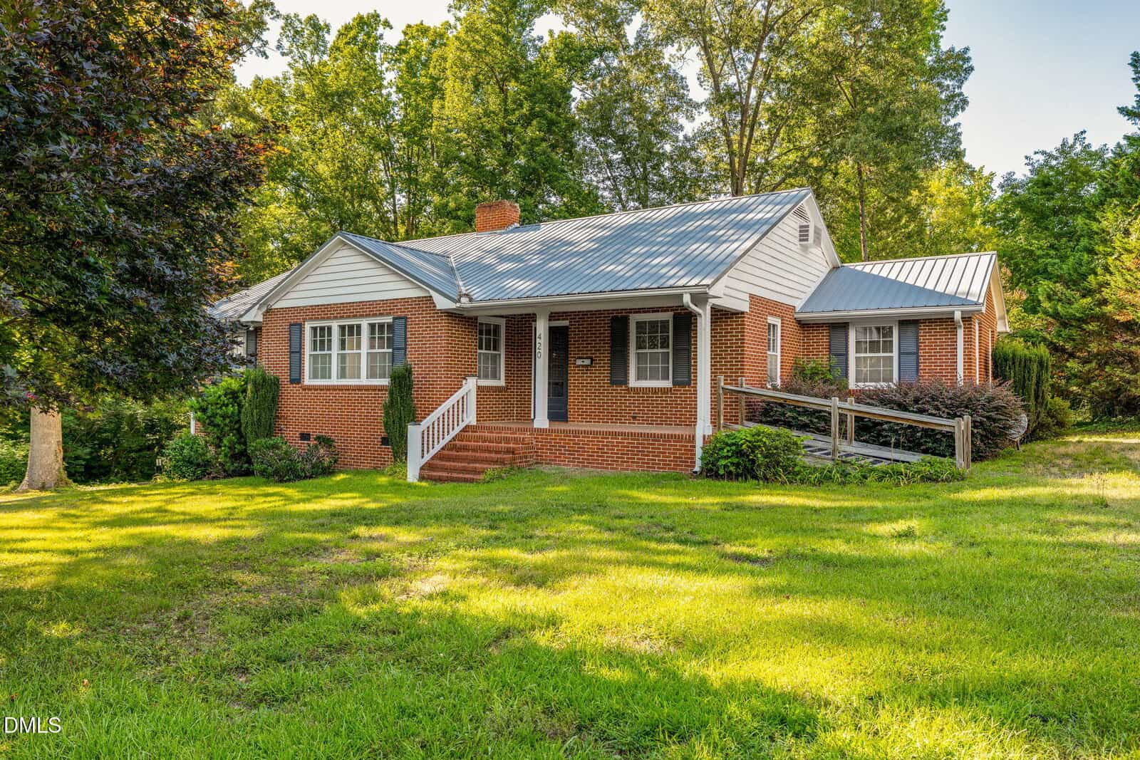 Single-story red brick house with a gray metal roof, surrounded by green trees and grass. The front porch has steps and a ramp for accessibility, and bushes line the walkway.