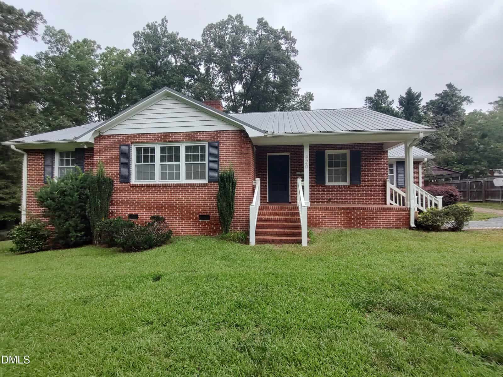 A one-story red brick house with white trim, a metal roof, and two sets of steps leading to separate entrances sits at 420 Rodwell Street in Warrenton, surrounded by green grass and trees on a cloudy day.