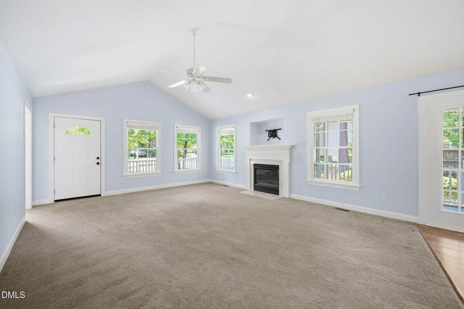 A spacious, light-filled living room with beige carpet, pale blue walls, large windows, a white front door, a ceiling fan, and a white fireplace with a mantel under a vaulted ceiling.