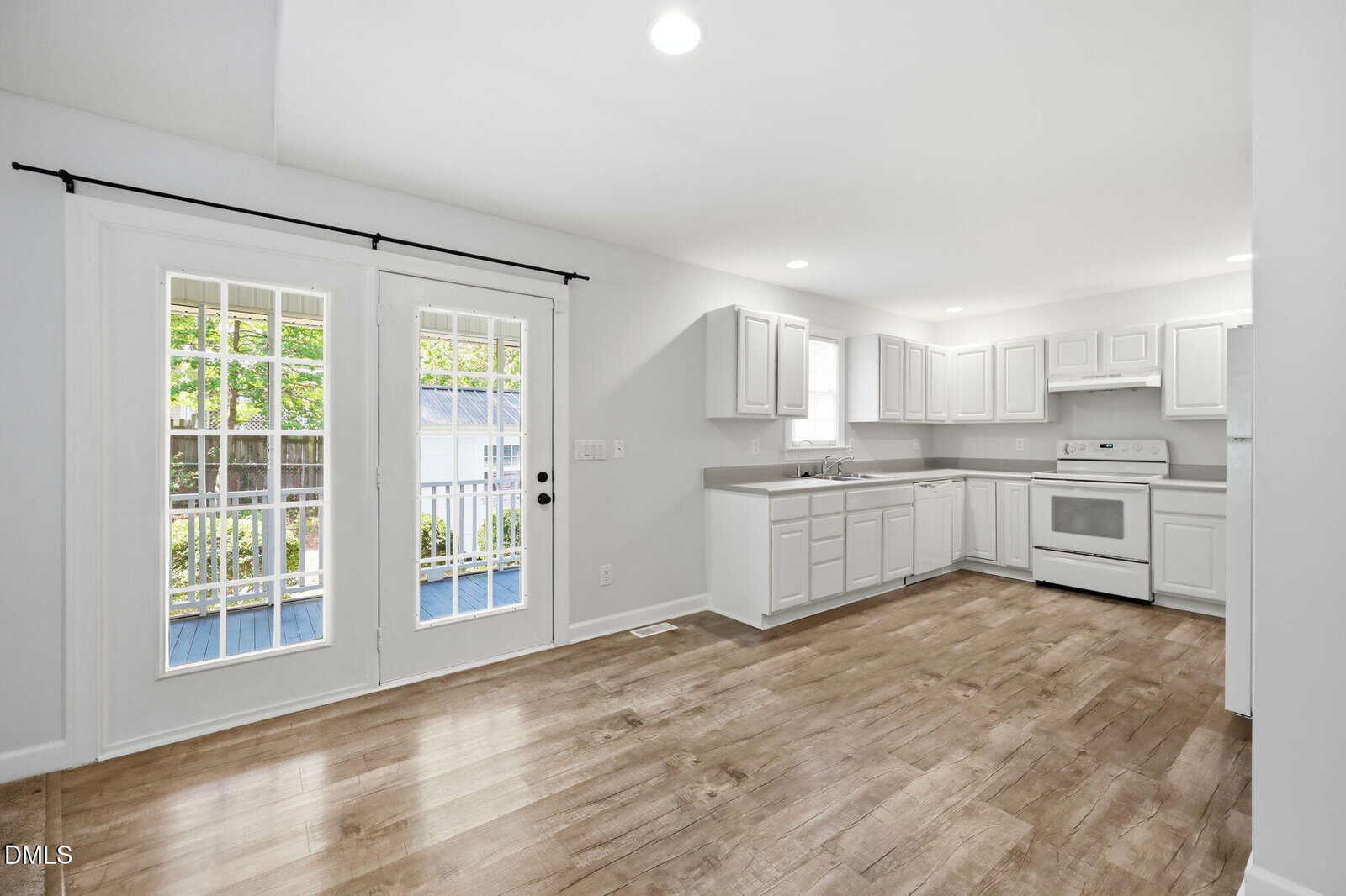 Bright, modern kitchen with white cabinets and appliances, wood-style flooring, and double glass doors leading to an outdoor area with a porch and greenery visible through the windows.