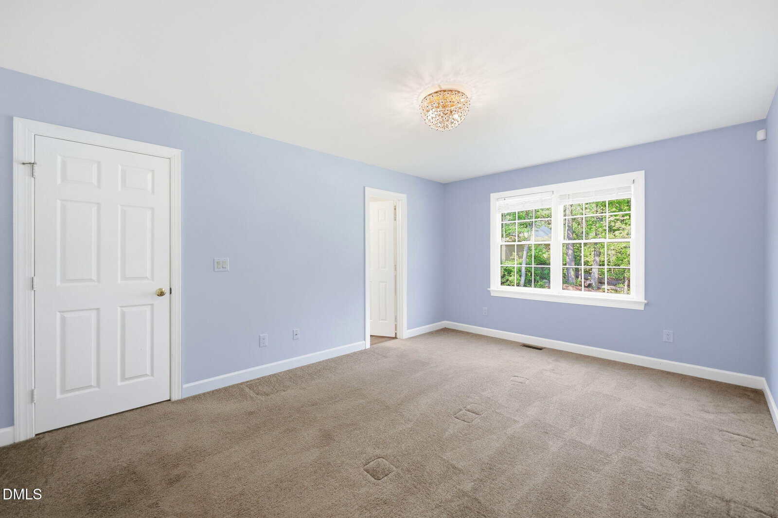 A bright, empty bedroom with light blue walls, beige carpet, a window letting in natural light, a white door, and a ceiling light fixture.