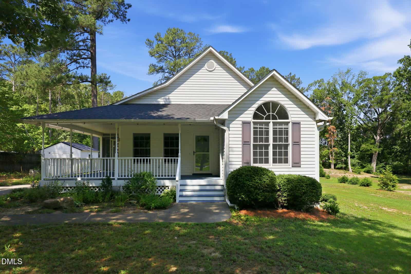 A white house with a front porch, large window with shutters, steps leading to the door, surrounded by trees, grass, and bushes under a blue sky.