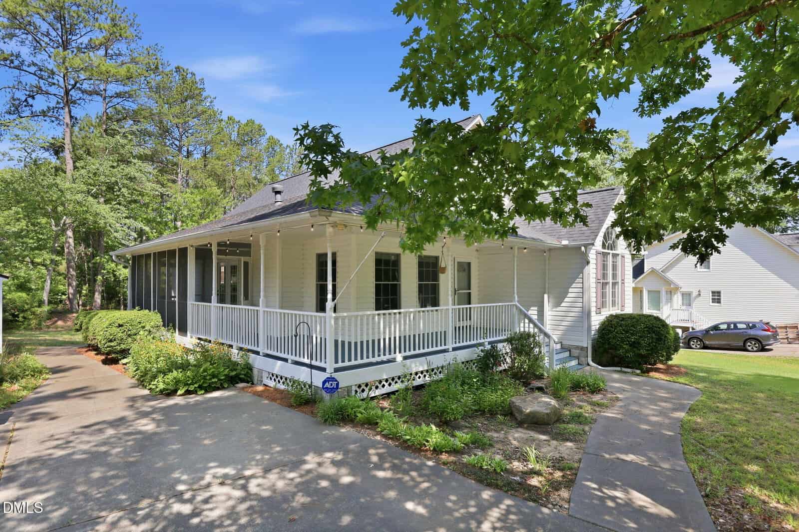 A white house with a large wraparound porch, surrounded by green trees and bushes, sits under a clear blue sky. The driveway curves beside the porch, and well-kept landscaping borders the walkway.