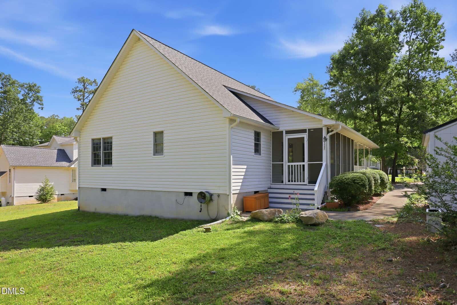 A single-story white house with a screened-in porch, gray roof, and green lawn. Trees and shrubs surround the home, and a small walkway leads to the porch under a clear blue sky.