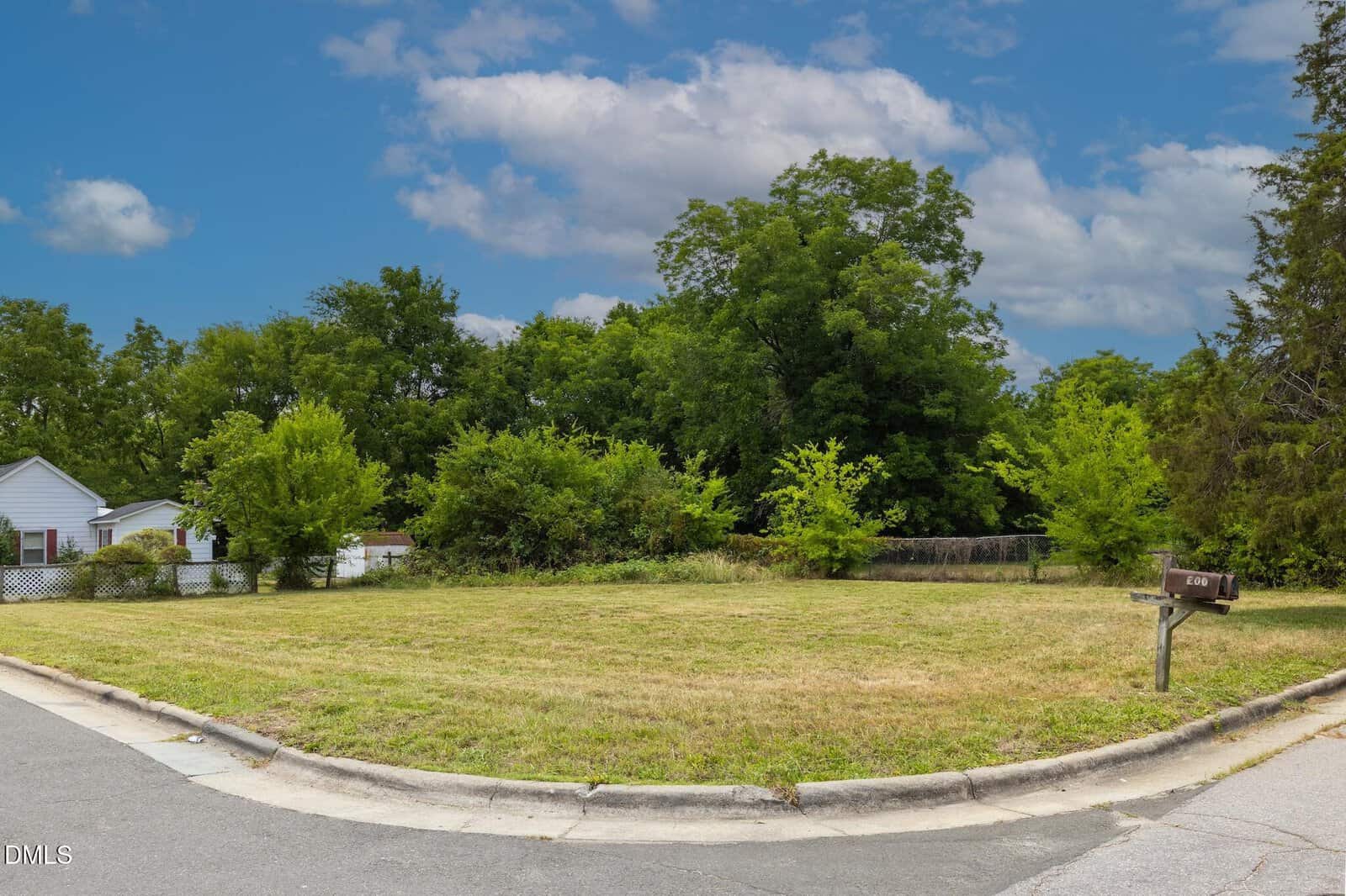 A corner lot with trimmed grass, some small trees, and dense greenery in the background. A mailbox stands near the curb, and a white house with a picket fence is partially visible on the left side.