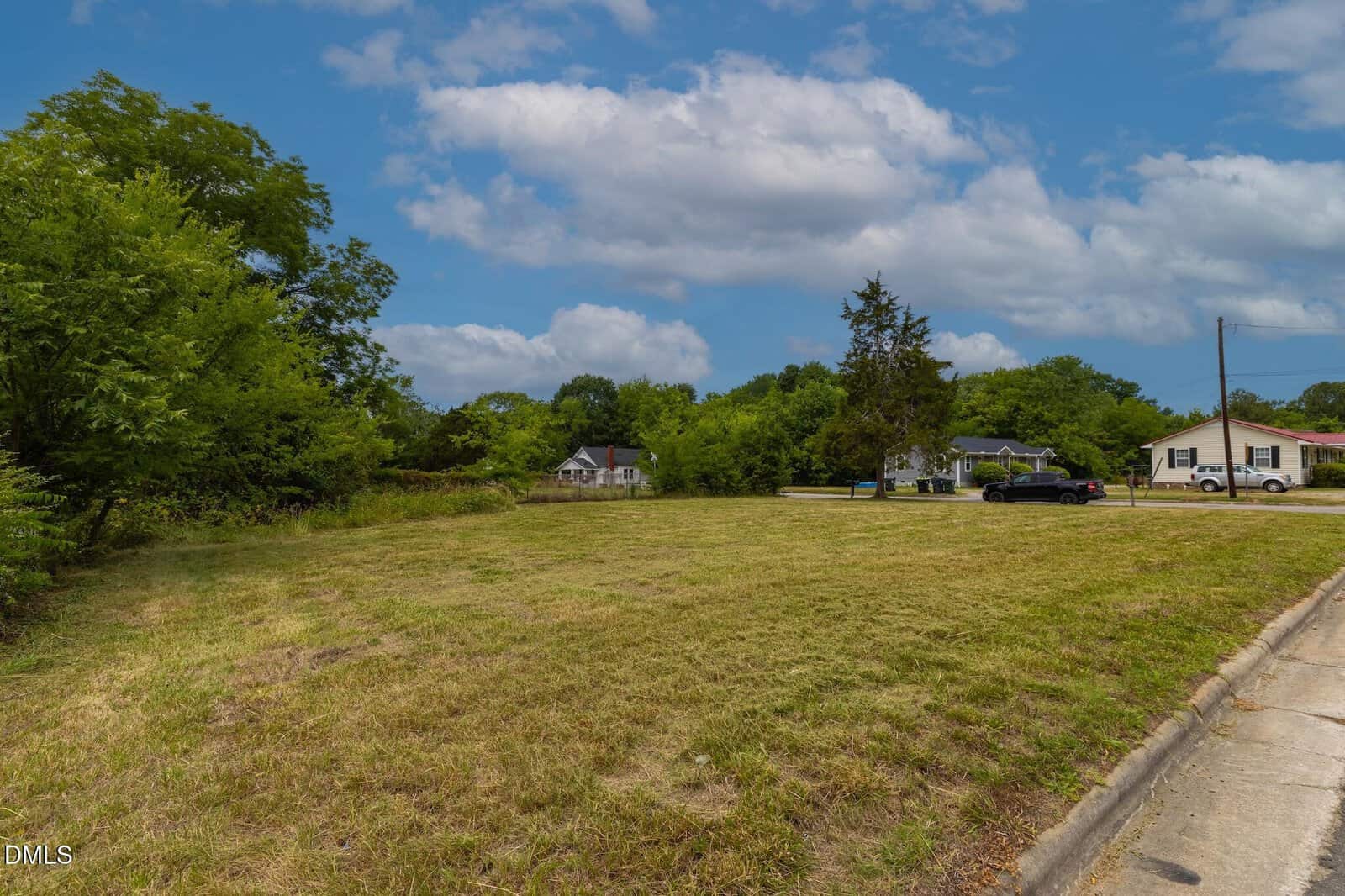 A grassy empty lot bordered by trees, with a few houses, cars, and a sidewalk in the background under a partly cloudy sky.
