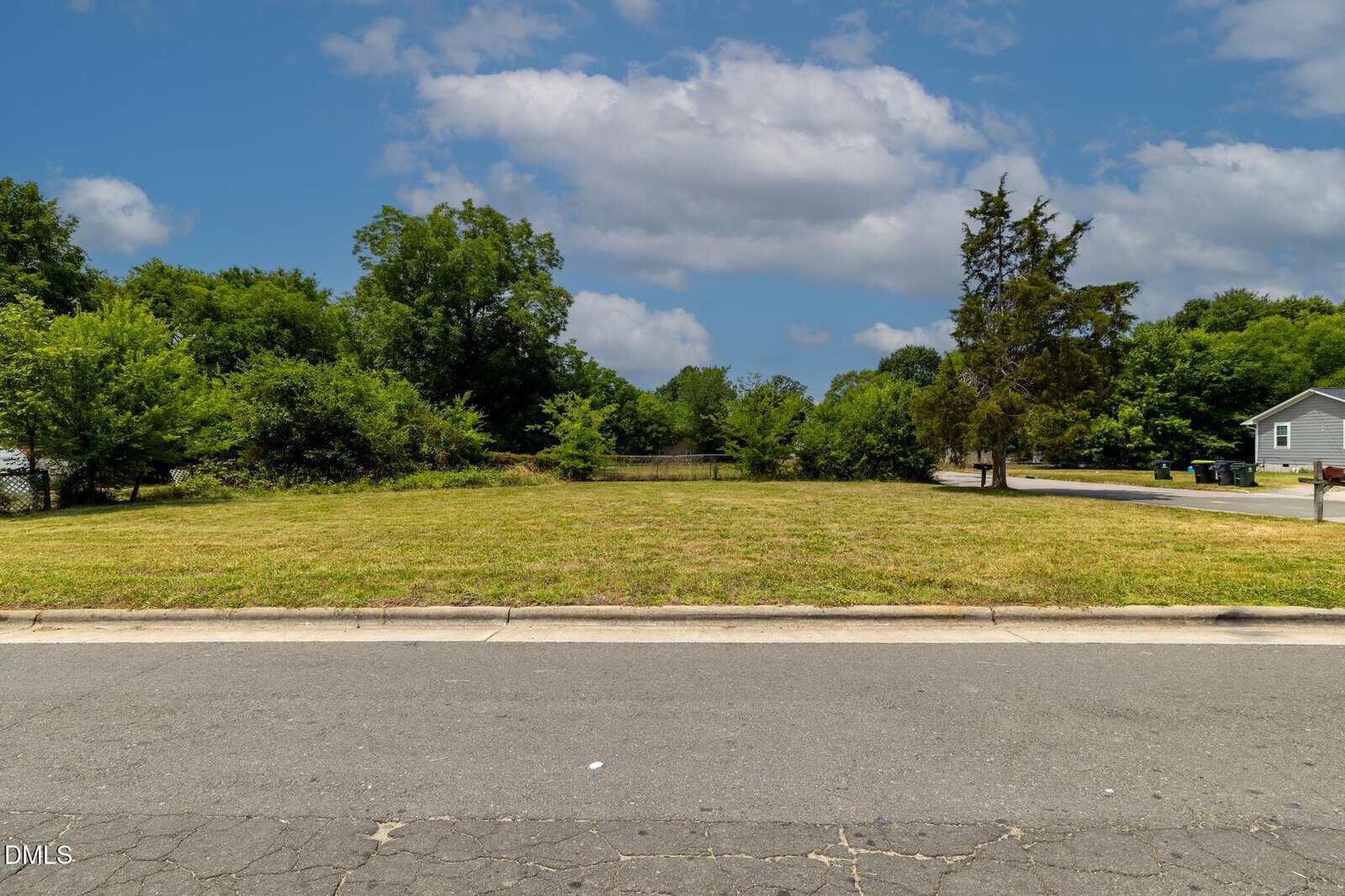 A vacant grassy lot bordered by trees, with a paved road in front. There is a small house and trash bins to the right, and a blue sky with scattered clouds overhead.