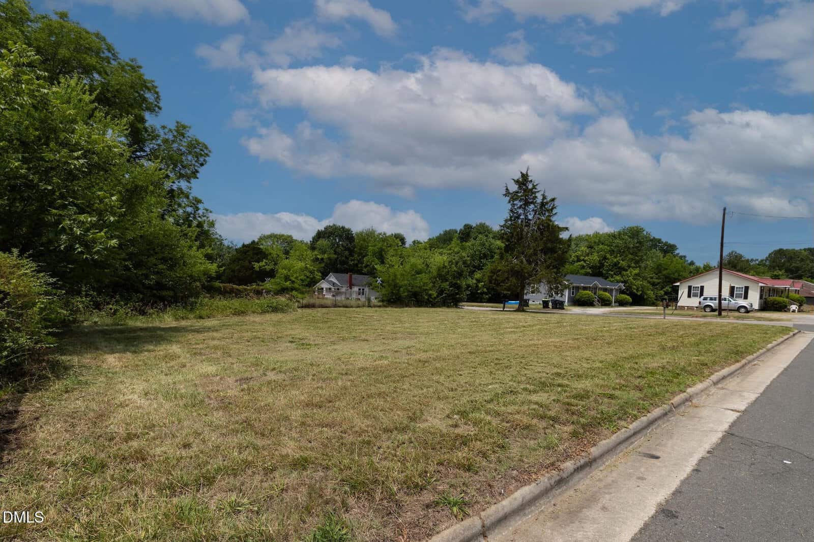A grassy vacant lot next to a street with a few houses in the background, surrounded by trees under a partly cloudy sky.