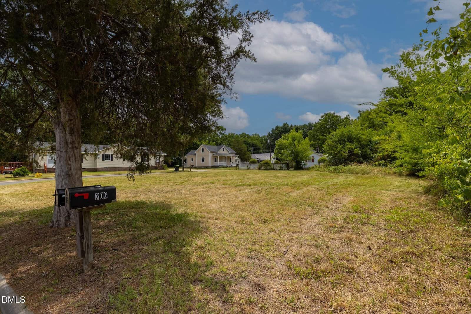 A grassy, empty lot with a mailbox labeled 2016 in the foreground, bordered by trees and bushes. In the background, a few houses are visible under a partly cloudy sky.
