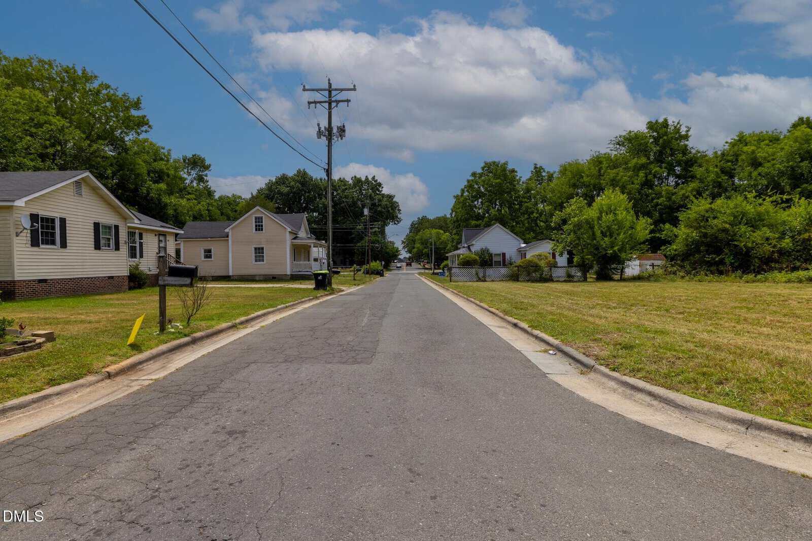 A quiet suburban street lined with single-story houses, green lawns, and trees under a partly cloudy sky. There are mailboxes and power lines along the road.