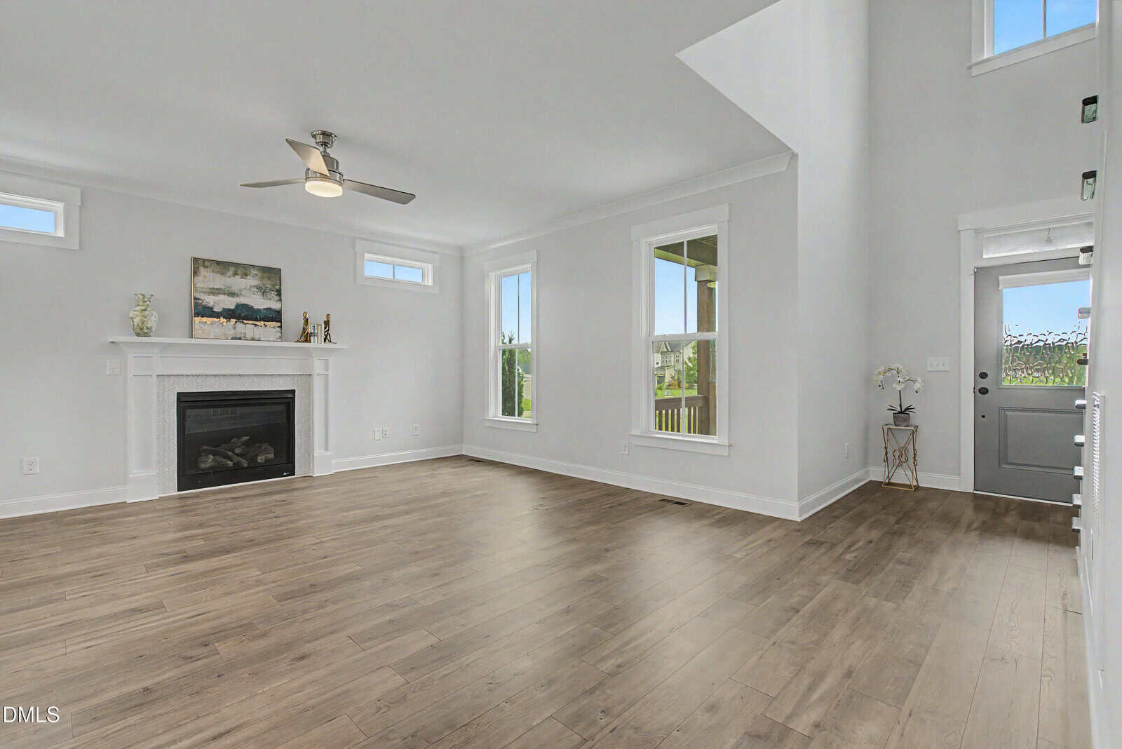 Spacious living room with light gray walls, wood flooring, a ceiling fan, white-trimmed windows, a white fireplace with decor, and a gray front door with glass panels. Natural light fills the room from multiple windows.