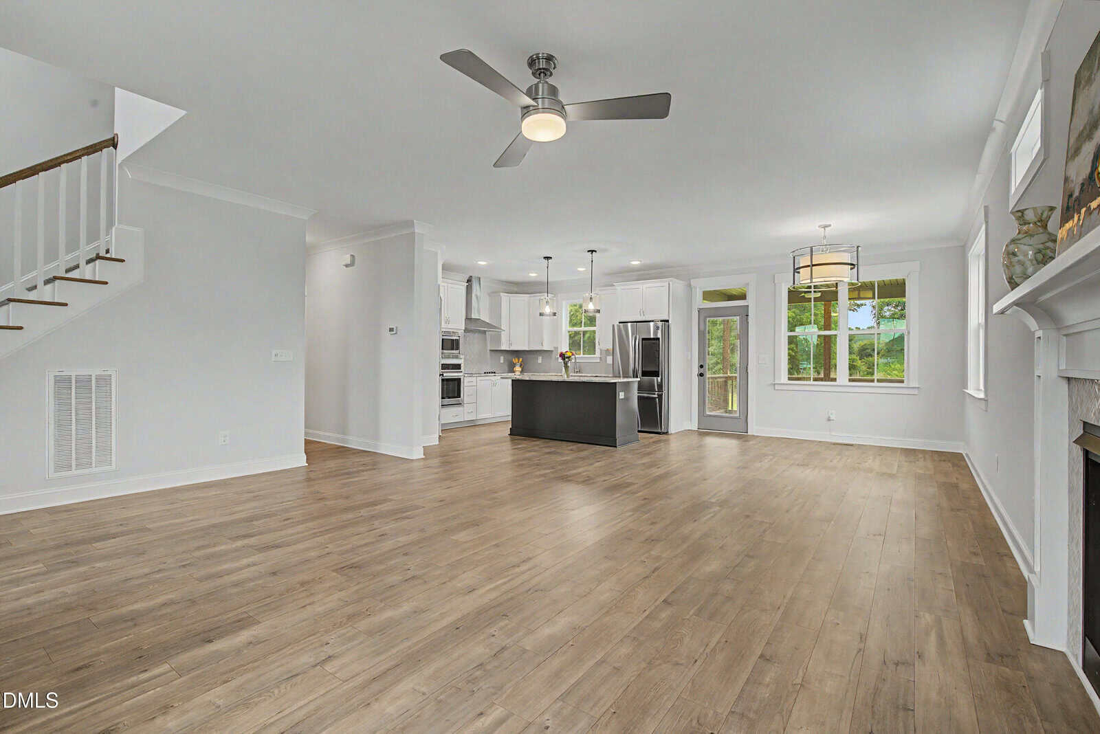 Modern open-concept living area with wood flooring, white walls, ceiling fans, a fireplace, and a kitchen with stainless steel appliances, island, and pendant lights. Large windows bring in natural light.