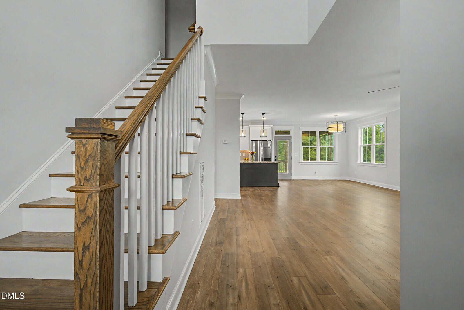 A modern, open-concept home interior with wood flooring, white walls, a staircase with wooden railing, and a view of the kitchen and living area with large windows and pendant lights.