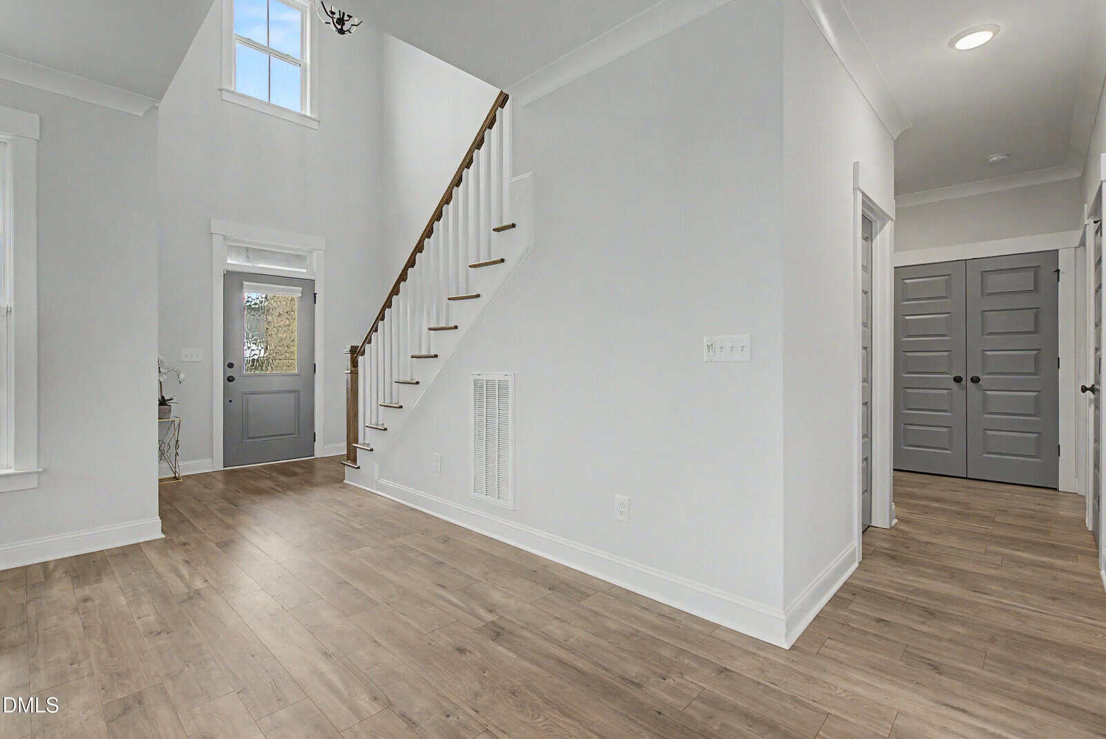 A modern, bright entryway features light wood flooring, white walls, a staircase with wooden handrails, gray doors, and a tall window above the front door.