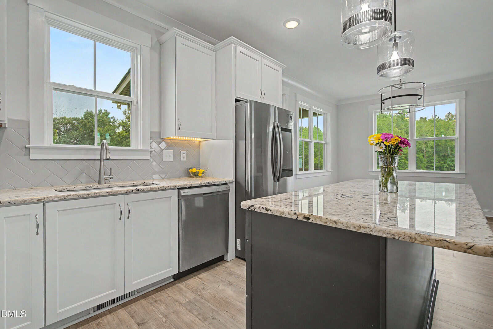 Modern kitchen with white cabinets, stainless steel appliances, a marble island with a vase of flowers, pendant lights, large windows, and light wood flooring. Green trees are visible through the windows.