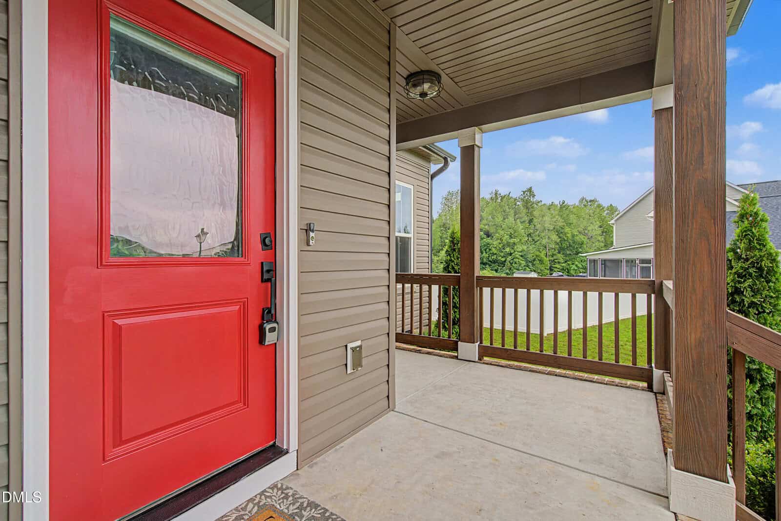 A close-up view of a red front door on a covered porch with beige siding, wood railing, and a concrete floor, overlooking a green yard and neighboring houses under a blue sky.