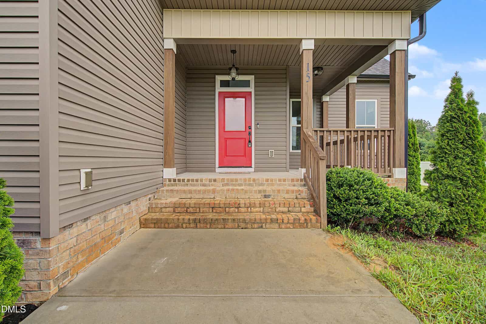 A concrete walkway leads to a small porch with brick steps, a red front door, tan siding, and wooden railings. Bushes and greenery line the edge of the porch.