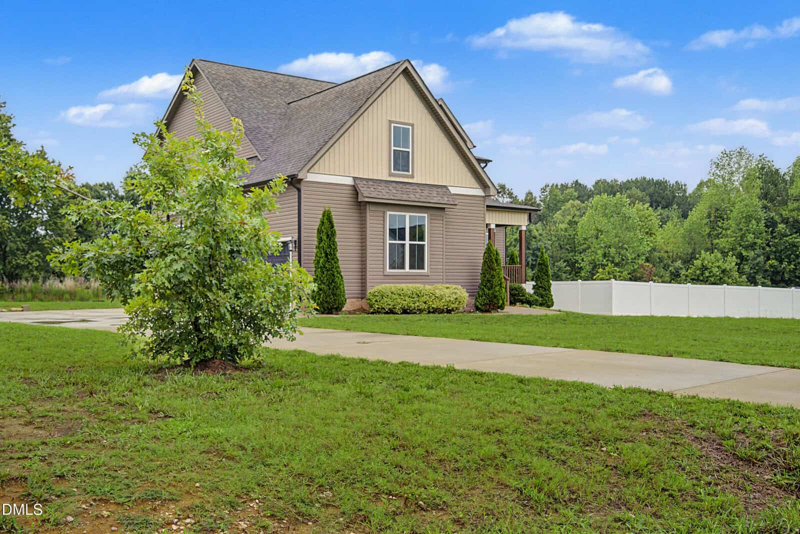 A modern beige and tan house with a steep roof, surrounded by green grass, small trees, and bushes, with a concrete driveway and a white fence in the background under a blue sky.