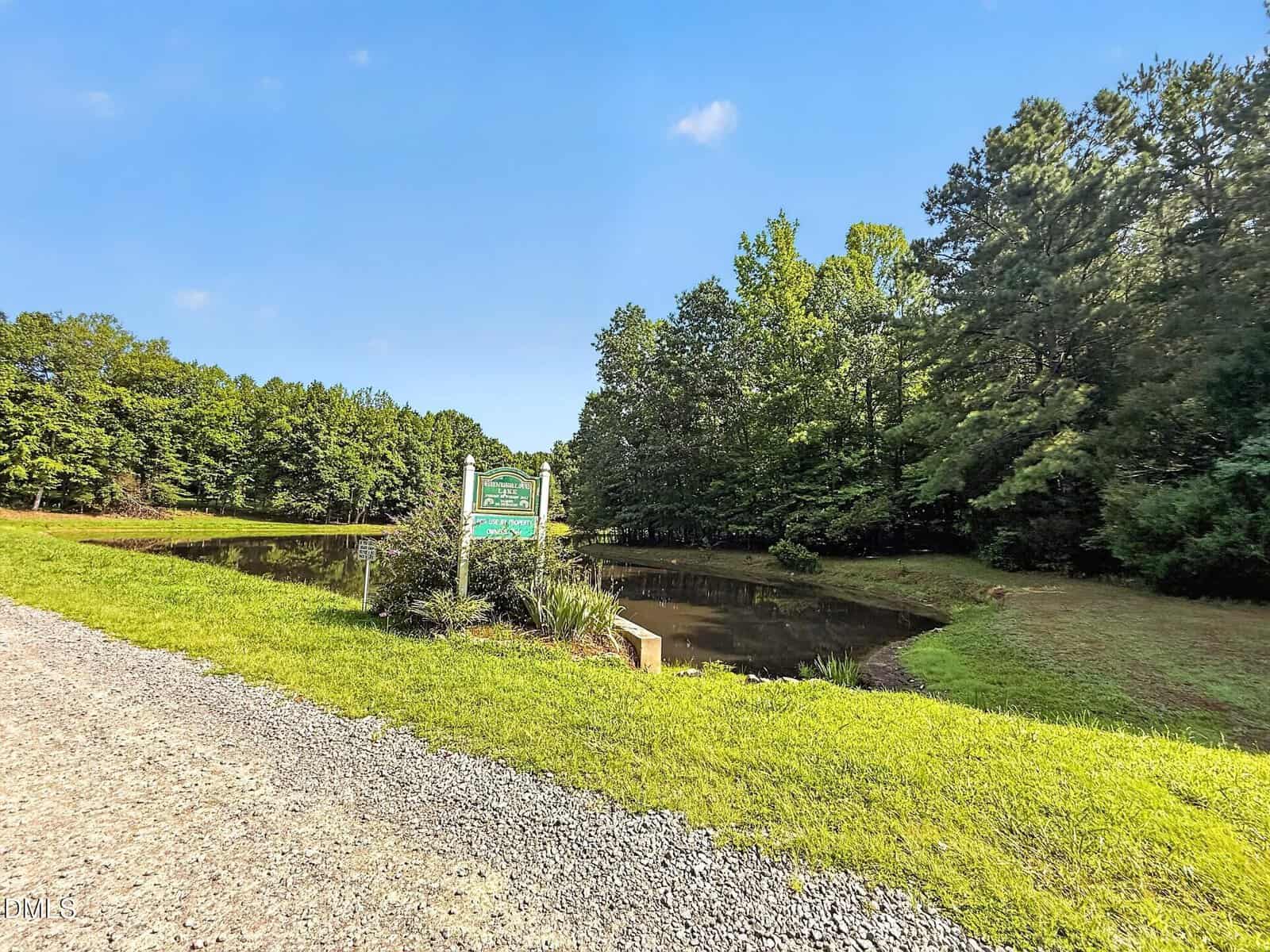 A small pond surrounded by trees sits beside a gravel road under a clear blue sky. A green sign on the grass marks the pond’s location.