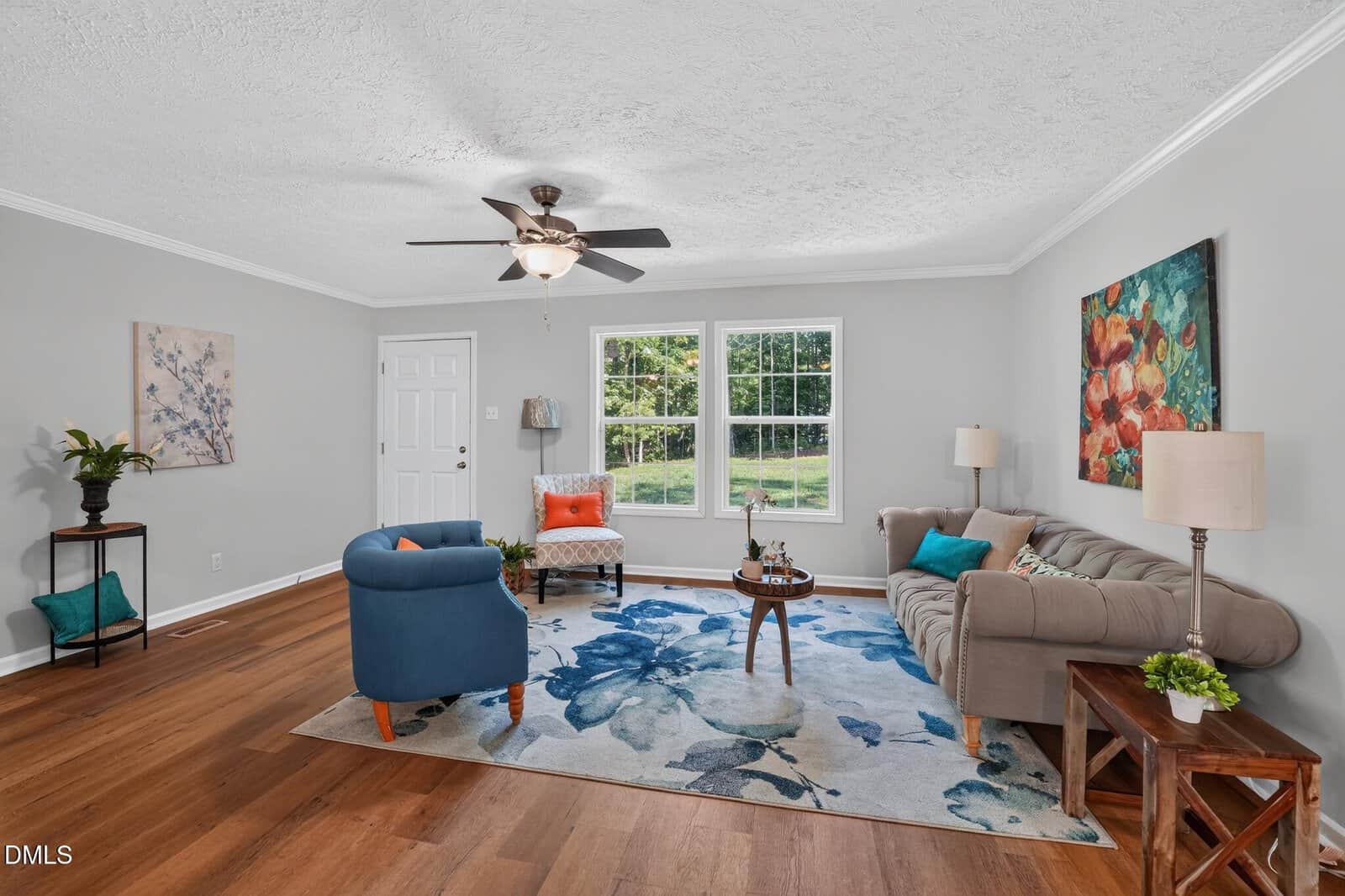 Bright living room with wood floors, a blue floral rug, modern furniture, large window with outdoor view, ceiling fan, colorful artwork, and decorative accents like plants and lamps.